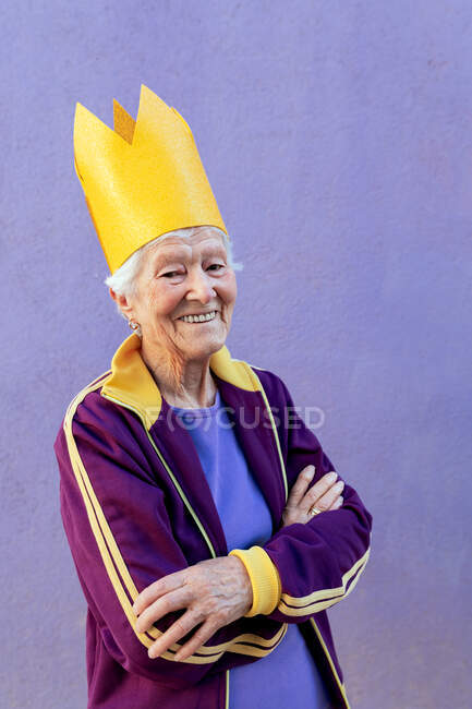 Уверенная в себе пожилая спортсменка в спортивной одежде и декоративной короне смотрит на камеру со сложенными руками на фиолетовом фоне — стоковое фото
