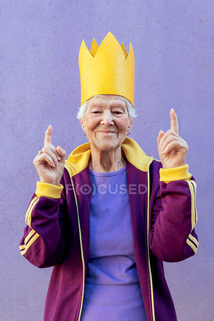 Contenido atleta femenina senior en ropa deportiva y corona de papel apuntando hacia arriba con los dedos mientras mira la cámara sobre fondo violeta - foto de stock