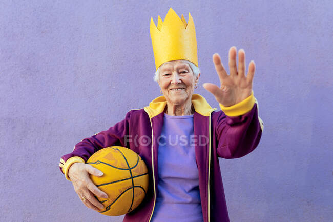 Fröhliche Seniorin in Sportbekleidung mit Basketball blickt in die Kamera und demonstriert Kontrollgeste auf lila Hintergrund — Stockfoto