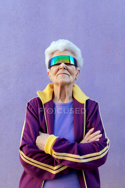Atleta anciana con brazos cruzados y canas en ropa deportiva y venda en fondo violeta - foto de stock
