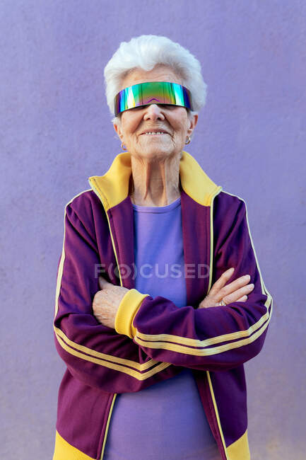 Alegre atleta anciana con los brazos cruzados y pelo gris en ropa deportiva y venda en el fondo violeta - foto de stock