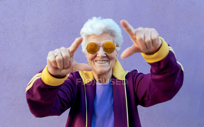 Smiling senior female athlete in modern sunglasses demonstrating frame gesture with outstretched arms on violet background — Stock Photo
