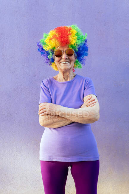 Content senior female athlete in modern sunglasses and bright wig standing with folded arms on purple background — Stock Photo