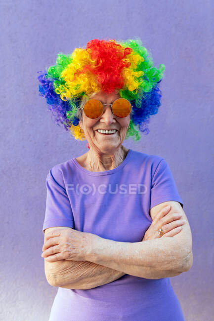 Zufriedene Seniorin mit moderner Sonnenbrille und heller Perücke mit verschränkten Armen auf lila Hintergrund — Stockfoto