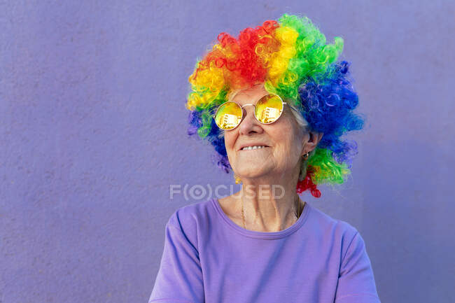 Contenuto atleta donna anziana in occhiali da sole moderni e parrucca luminosa in piedi su sfondo viola — Foto stock