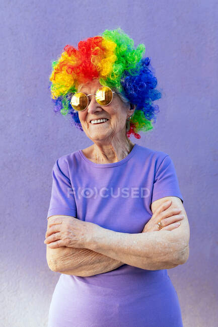 Athlète féminine senior en lunettes de soleil modernes et perruque lumineuse debout avec les bras croisés sur fond violet — Photo de stock
