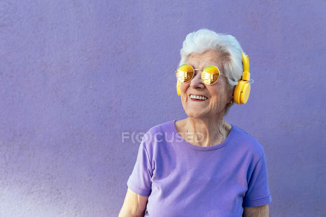 Mujer mayor alegre en camiseta y gafas de sol modernas escuchando la canción de auriculares inalámbricos sobre fondo púrpura - foto de stock