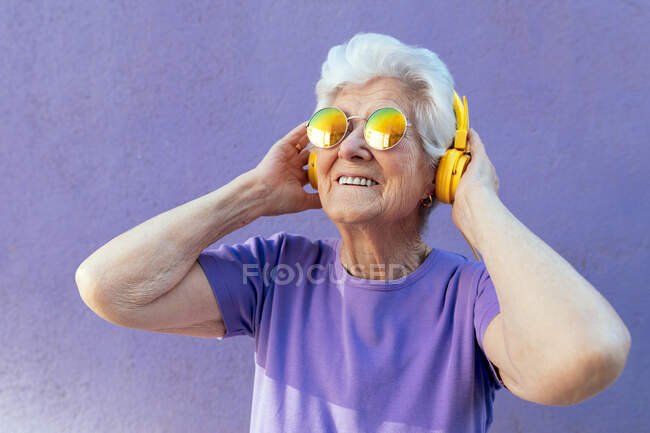 Allegro anziano donna in t-shirt e occhiali da sole moderni ascoltando la canzone da auricolare wireless su sfondo viola — Foto stock