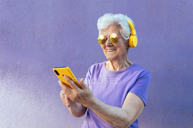 Весела літня жінка в сучасних сонцезахисних окулярах та навушниках слухати музику під час серфінгу в Інтернеті на мобільному телефоні на фіолетовому фоні — стокове фото