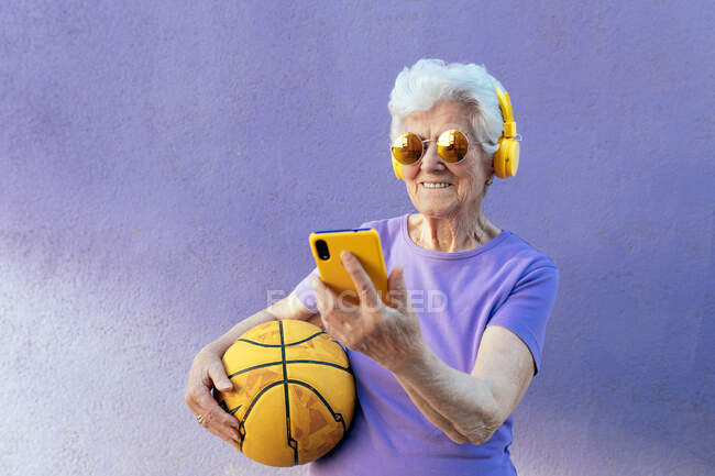 Mujer alegre de edad avanzada en gafas de sol modernas y auriculares escuchando música mientras navega por Internet en el teléfono celular sobre fondo violeta - foto de stock
