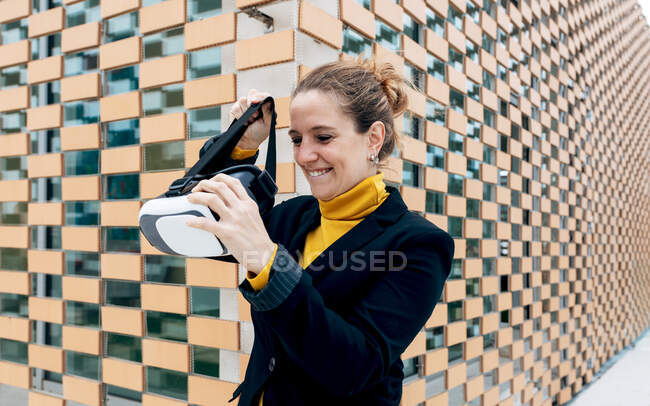 Mujer adulta sonriente con un atuendo elegante parada cerca de la pared del edificio con gafas VR y mirando hacia otro lado a la luz del día - foto de stock