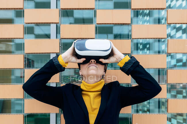 Femme en veste et col roulé à l'aide de lunettes VR près des fenêtres du bâtiment en plein jour — Photo de stock