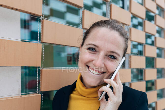 Позитивна доросла жінка в стильному вбранні, дивлячись на камеру, розмовляючи на смартфоні біля стіни будівлі вдень — стокове фото