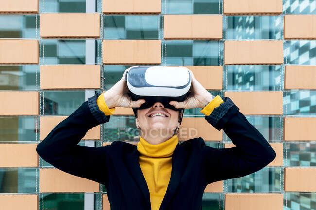 Femme souriante en veste et col roulé utilisant des lunettes VR près des fenêtres du bâtiment à la lumière du jour — Photo de stock