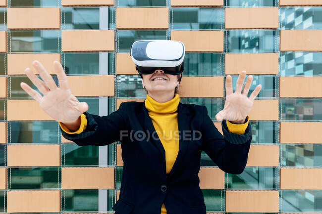 Femme souriante en veste et col roulé utilisant des lunettes VR près des fenêtres du bâtiment à la lumière du jour — Photo de stock