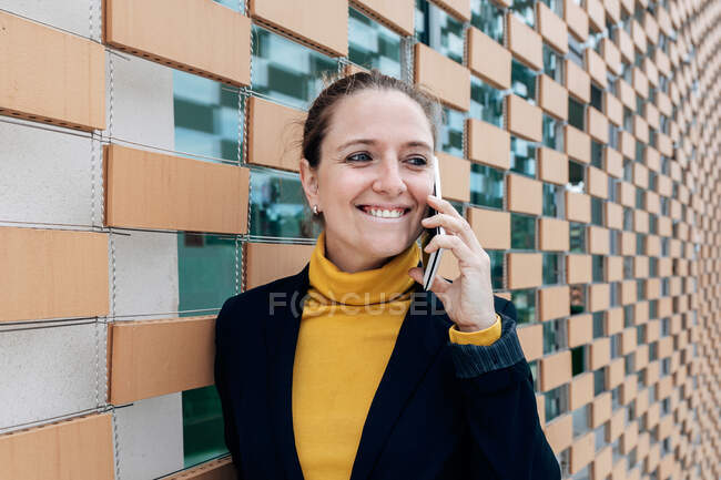 Positive erwachsene Frau in stylischem Outfit schaut weg, während sie tagsüber am Smartphone in der Nähe der Hauswand spricht — Stockfoto