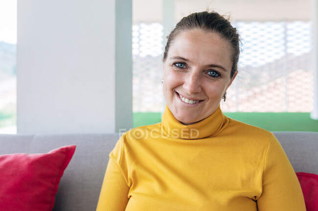 Glückliche erwachsene Frau in lässiger Kleidung sitzt auf der Couch mit Kissen und blickt in die Kamera im hellen Haus — Stockfoto