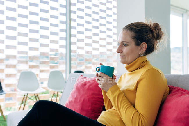 Thoughtful adult female in casual outfit sitting on couch with pillows with mug with drink in light room while looking away near windows and chairs — Stock Photo