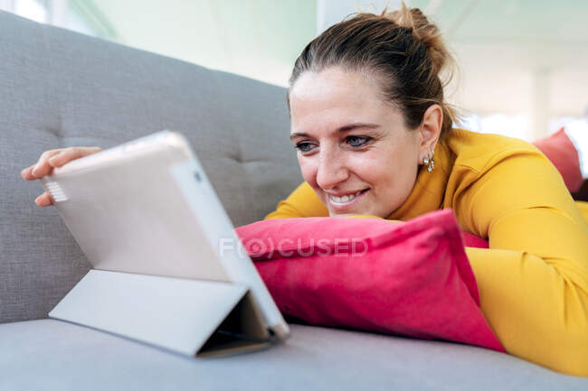 Mujer adulta positiva en ropa casual acostada en el sofá con almohada mientras navega en la tableta en la sala de estar ligera - foto de stock