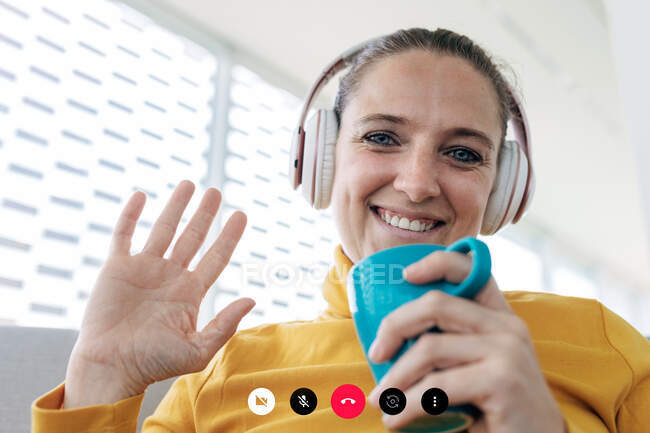 Low angle of happy adult female in casual clothes with headphones and cup of drink having video chat in light room while waving hand and looking at camera — Stock Photo