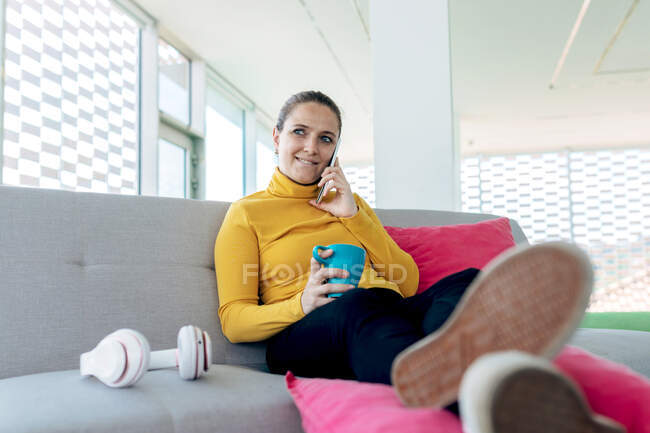 Positive adult female in casual clothes sitting on couch with mug with drink while talking on smartphone near headphones in light living room — Stock Photo