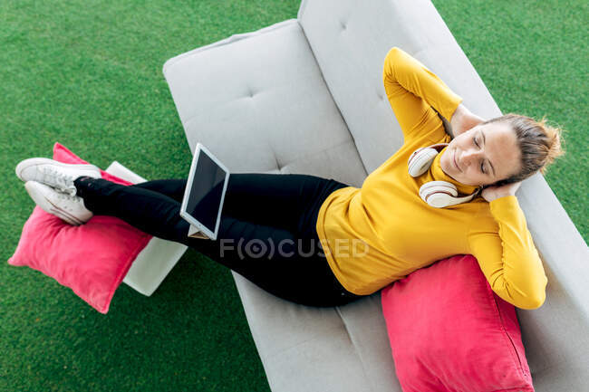 Dall'alto della femmina adulta positiva in abito casual con cuffie e tablet seduta sul divano con cuscini in studio leggero — Foto stock