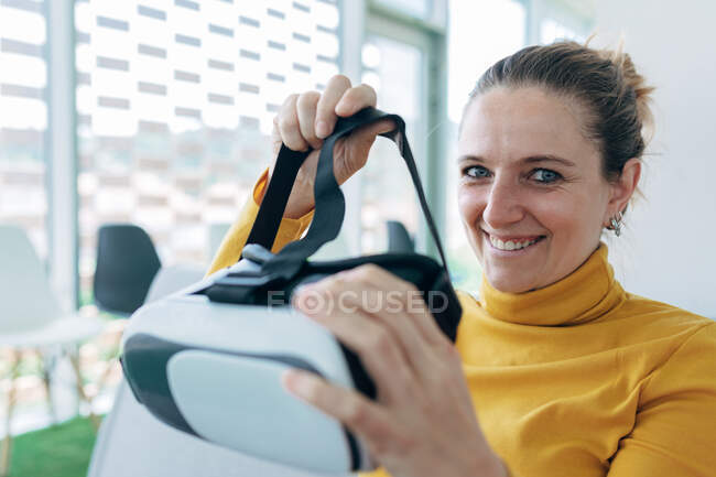Female in casual outfit sitting on sofa and using VR goggles near windows in light building — Stock Photo