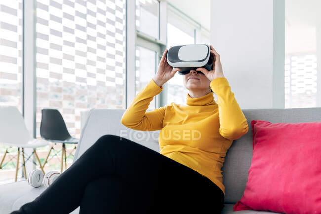 Женщина в повседневной одежде сидит на диване с подушками, используя защитные очки возле наушников и окон в светлом здании — стоковое фото