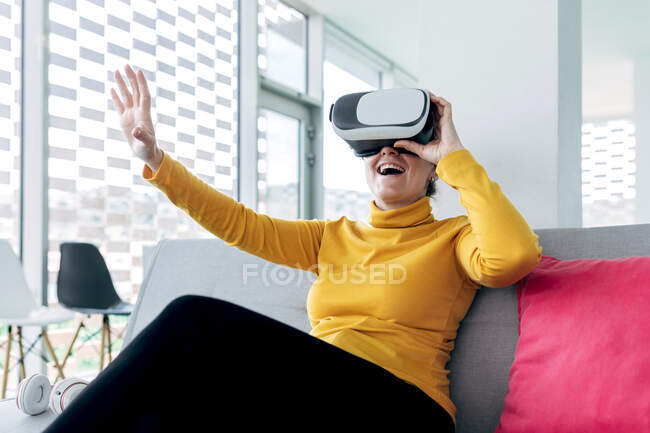 Feminino em roupa casual sentado no sofá com travesseiros enquanto usa óculos VR perto de fones de ouvido e janelas no prédio leve — Fotografia de Stock