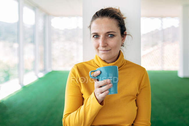 Positive erwachsene Frau in lässigem Outfit mit Kaffeebecher im hellen Raum mit Blick auf Kamera in der Nähe von Fenstern und Säule — Stockfoto