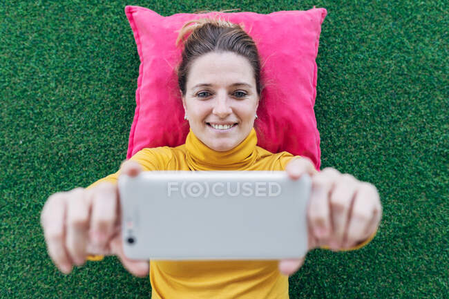 Зверху позитивна доросла жінка в повсякденному одязі лежить на подушці на зеленому килимі під час перегляду цікавого відео на мобільному телефоні — стокове фото