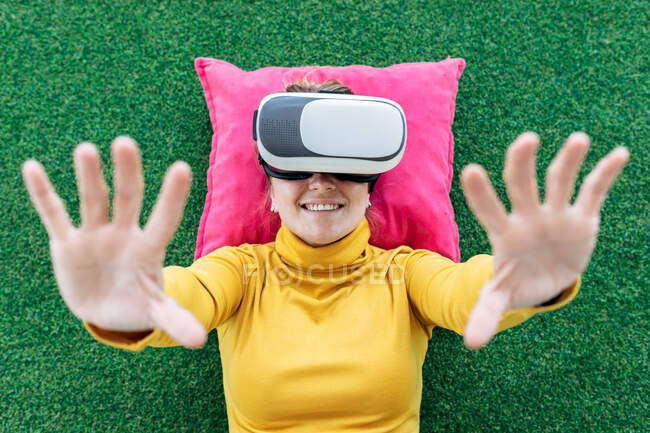Зверху усміхнена жінка в повсякденному одязі лежить на подушці на зеленому килимі, відчуваючи віртуальну реальність в гарнітурі VR з піднятими руками на камеру — стокове фото