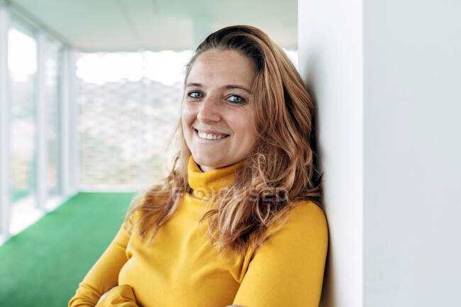 Mulher adulta feliz em blusa amarela olhando para a câmera enquanto estava perto da coluna no edifício de luz com janelas panorâmicas e piso verde — Fotografia de Stock