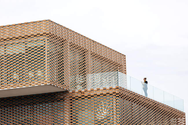 Von unten steht eine Frau in stilvollem Outfit auf dem Balkon eines modernen Gebäudes mit geometrischen Elementen an den Fenstern, während sie ein Tablet in der Nähe von Glasgeländern unter hellem Himmel benutzt — Stockfoto