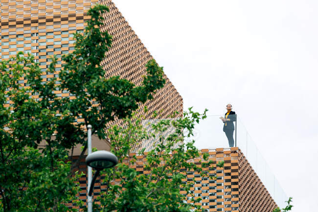 De dessous de femelle en tenue élégante debout sur le balcon du bâtiment moderne avec des éléments géométriques sur les fenêtres tout en utilisant une tablette près de balustrades en verre sous un ciel lumineux près des arbres verts — Photo de stock