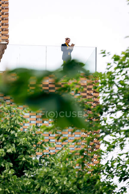 Von unten steht eine Frau in stilvollem Outfit auf dem Balkon eines modernen Gebäudes mit geometrischen Elementen an den Fenstern, während sie ein Tablet in der Nähe von Glasgeländern unter hellem Himmel in der Nähe grüner Bäume benutzt — Stockfoto