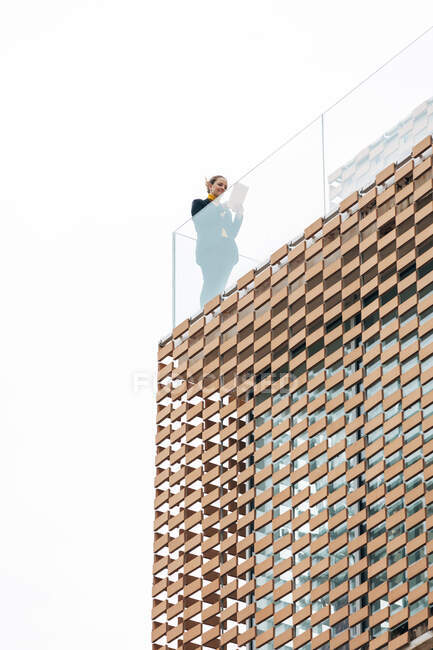 De baixo da fêmea na roupa elegante que está na varanda do edifício moderno com elementos geométricos em janelas ao usar a pastilha perto de grades de vidro abaixo do céu brilhante — Fotografia de Stock