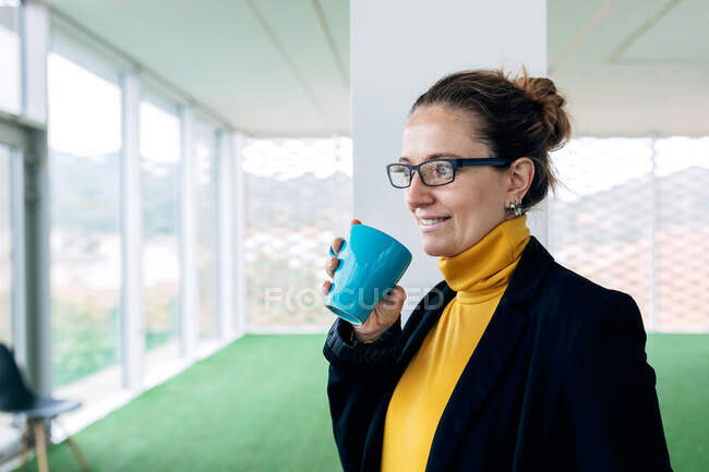 Mujer adulta positiva en traje elegante con taza con café en la sala de luz mirando lejos cerca de las ventanas y la columna - foto de stock
