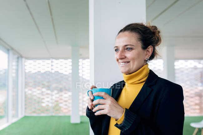 Mujer adulta positiva en traje elegante con taza con café en la sala de luz mirando lejos cerca de las ventanas y la columna - foto de stock