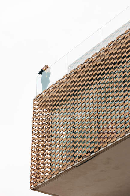 Von unten steht eine Frau in stilvollem Outfit auf dem Balkon eines modernen Gebäudes mit geometrischen Elementen an den Fenstern, während sie ein Tablet in der Nähe von Glasgeländern unter hellem Himmel benutzt — Stockfoto