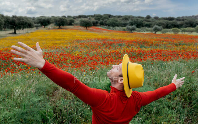 Vista posteriore di elegante maschio in abiti rossi e cappello giallo braccia tese in piedi con gli occhi chiusi in lussureggiante campo fiorito in una natura tranquilla — Foto stock