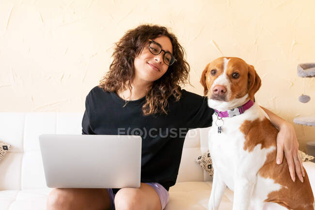 Молодая латиноамериканка гладит собаку и просматривает интернет на ноутбуке, проводя свободное время вместе в гостиной — стоковое фото
