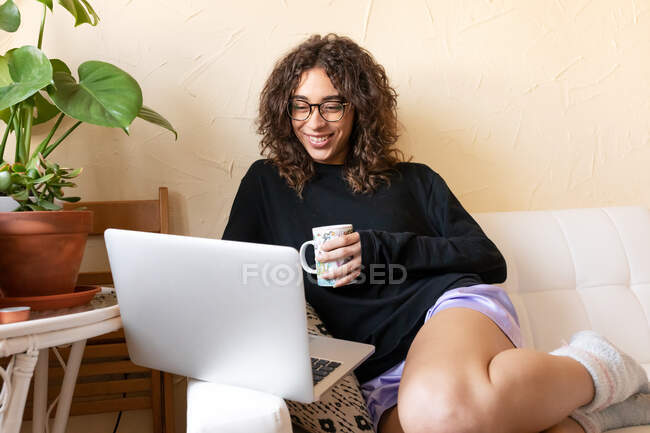 Молодая женщина в повседневной одежде и очках наслаждаясь горячим кофе вдумчиво сидя с ноутбуком на диване и охлаждения в одиночестве дома — стоковое фото