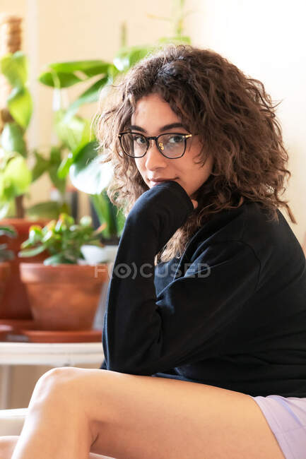 Vista lateral de una joven hispana de pelo rizado milenaria en ropa hogareña y anteojos mirando a la cámara mientras está sentada cerca de plantas en maceta en la sala de luz en casa - foto de stock
