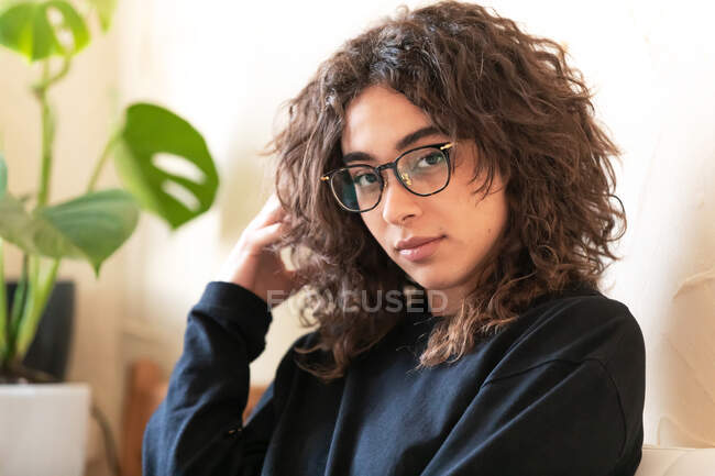 Seitenansicht der jungen lockigen hispanischen Millennial-Frau in heimeliger Kleidung und Brille, die in die Kamera schaut, während sie in der Nähe von Topfpflanzen in einem hellen Raum zu Hause sitzt — Stockfoto