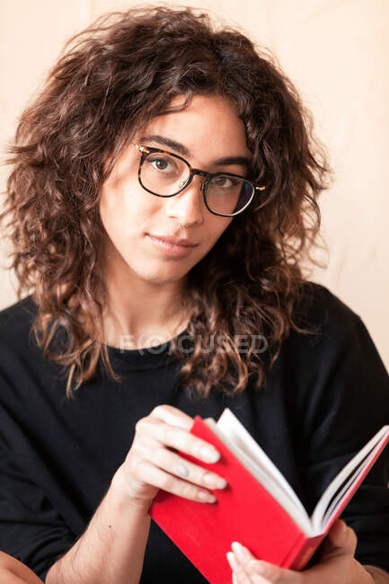 Giovane dai capelli ricci femmina ispanica in abiti casual e occhiali leggere il libro rosso e godersi la storia interessante durante il tempo libero a casa — Foto stock