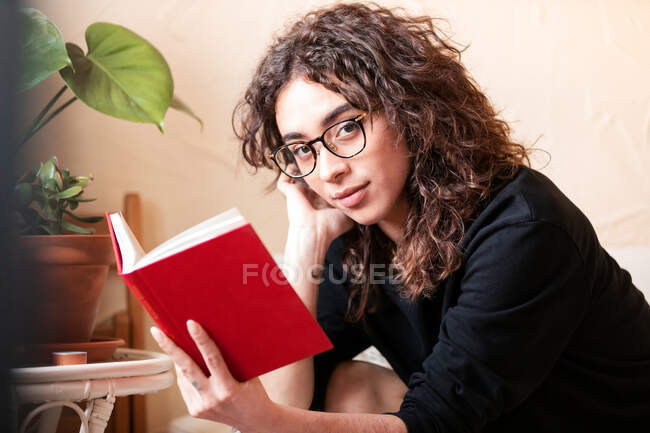 Jovem de cabelos encaracolados Mulher hispânica em roupas casuais e óculos lendo livro vermelho e desfrutando de história interessante durante o tempo livre em casa — Fotografia de Stock