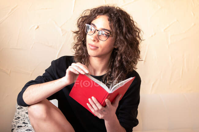 Giovane dai capelli ricci femmina ispanica in abiti casual e occhiali leggere il libro rosso e godersi la storia interessante durante il tempo libero a casa — Foto stock
