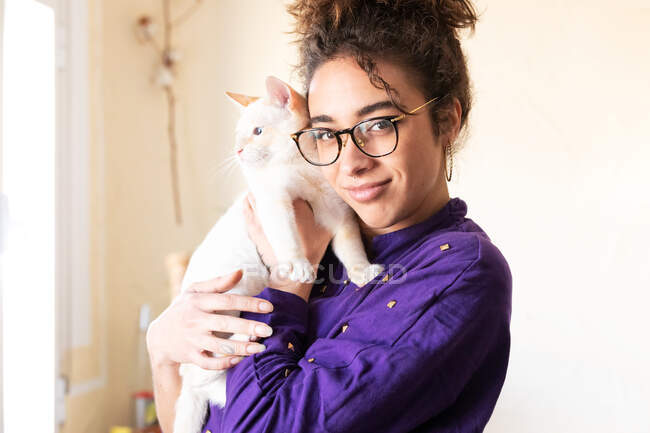 Happy young curly haired Hispanic female owner in eyeglasses cuddling cute domestic cat and looking at camera while resting together at home — Stock Photo