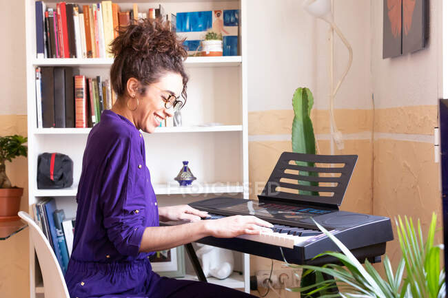 Seitenansicht einer fröhlichen jungen hispanischen Frau, die zu Hause E-Piano spielt, während sie musiziert — Stockfoto